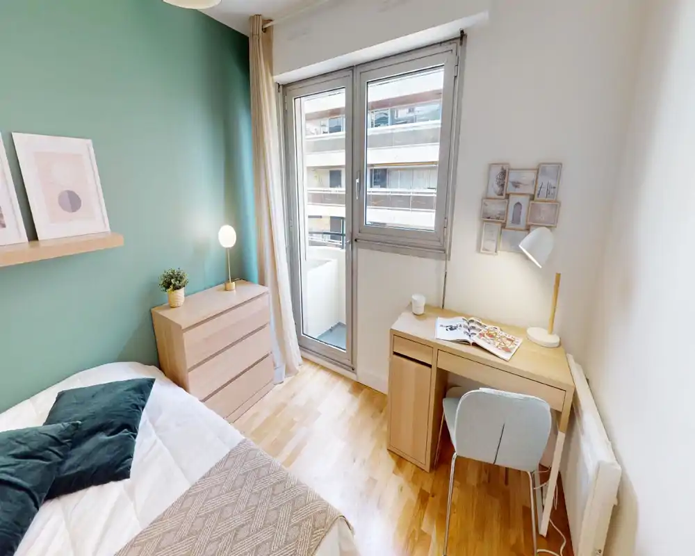 Bright, cozy bedroom with a light-wood chest of drawers, a comfortable bed with cushions, and a small desk by a large window leading to a balcony.