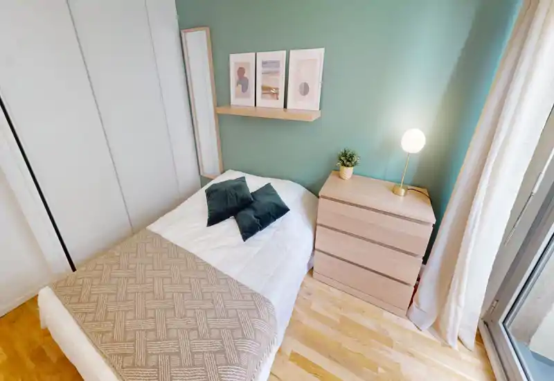 Bright, cozy single bedroom with a neatly made bed, bedside chest of drawers and a window letting in natural light. Calm green accent wall with framed artwork and a small lamp creates a pleasant, modern feel.