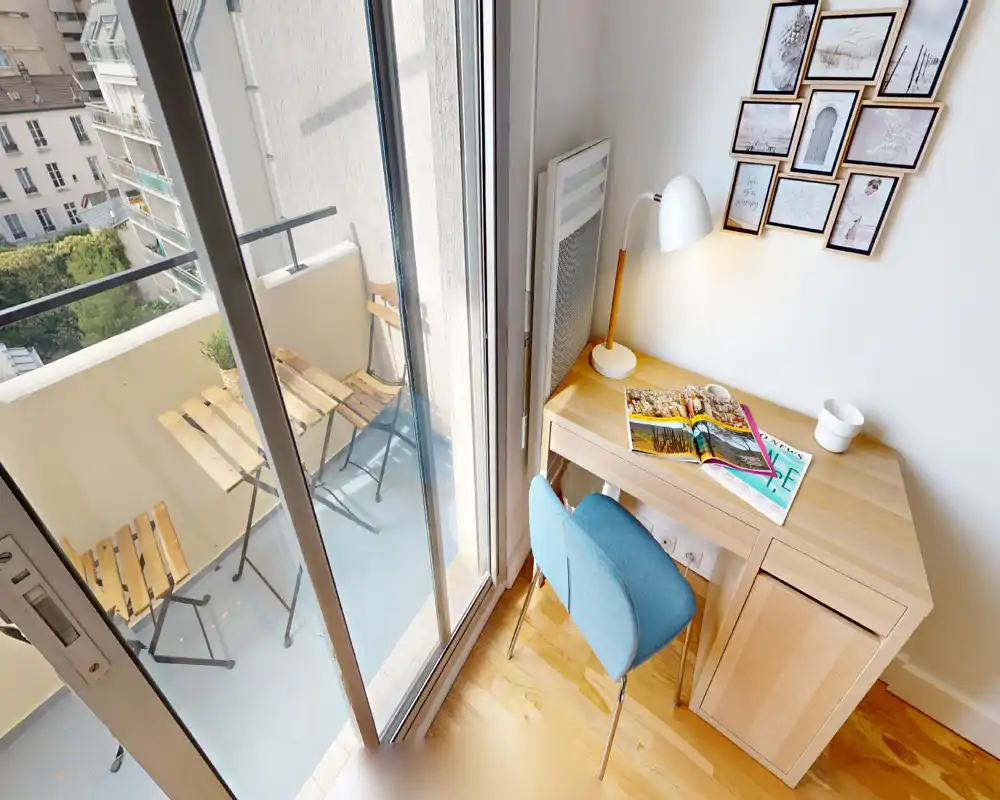 Coin salon lumineux et bien décoré avec un petit bureau et une chaise à côté d'une baie vitrée donnant sur un balcon compact équipé d'un ensemble bistrot en bois.