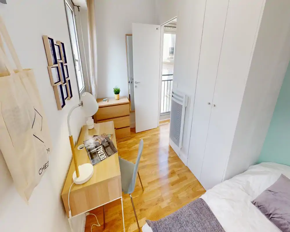 Chambre compacte et lumineuse avec lit simple partiellement visible, parquet et petit bureau près de la fenêtre — parfaite pour étudiant ou voyageur solo.
