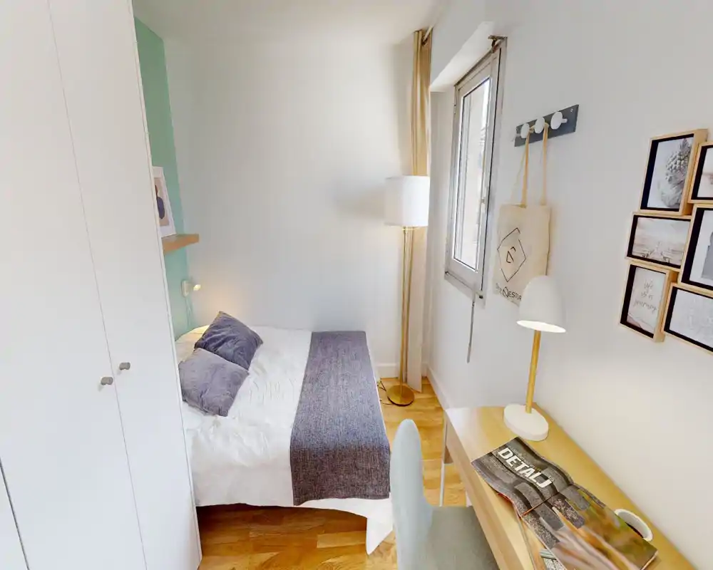 Chambre lumineuse et compacte avec un lit double en linge blanc et un jeté gris, une lampe sur pied près de la fenêtre et un bureau en bois avec lampe de lecture — idéale pour une location cosy et fonctionnelle.