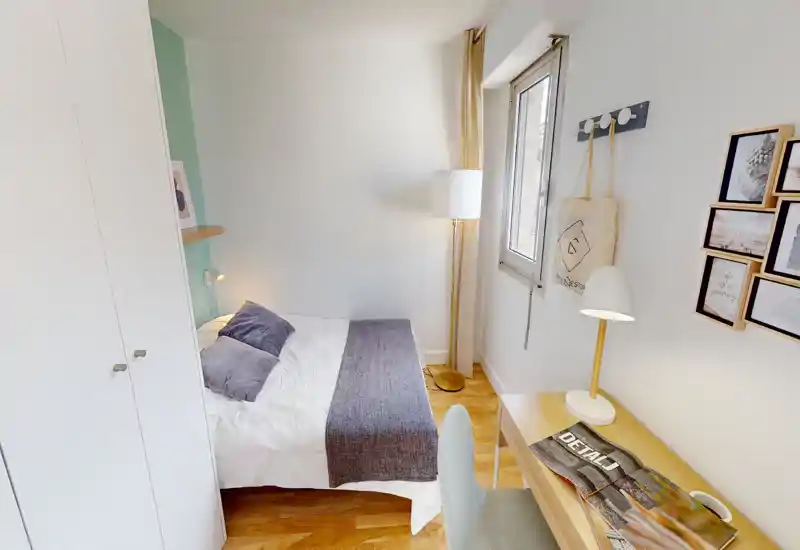 Bright, compact bedroom with a double bed dressed in white linens and a grey runner, a floor lamp by the window and a wooden desk with a reading lamp — ideal for a cozy, functional rental room.