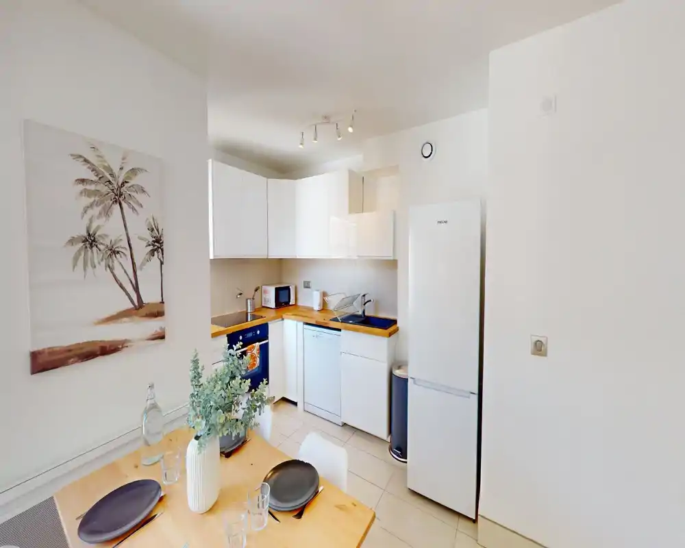 Compact, modern kitchen with white cabinets, wooden countertops, fridge, dishwasher and a small dining table set for two — bright and neatly styled.