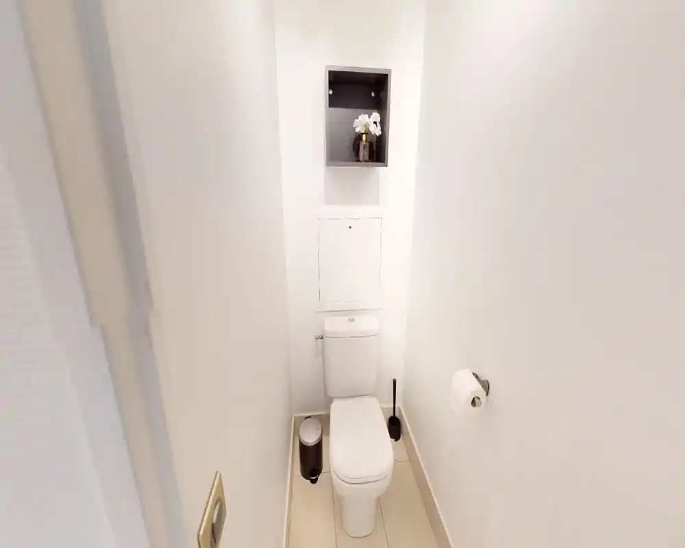 Compact, well-lit bathroom with a modern toilet, toilet brush and small waste bin; white walls and a recessed shelf with decorative flowers.