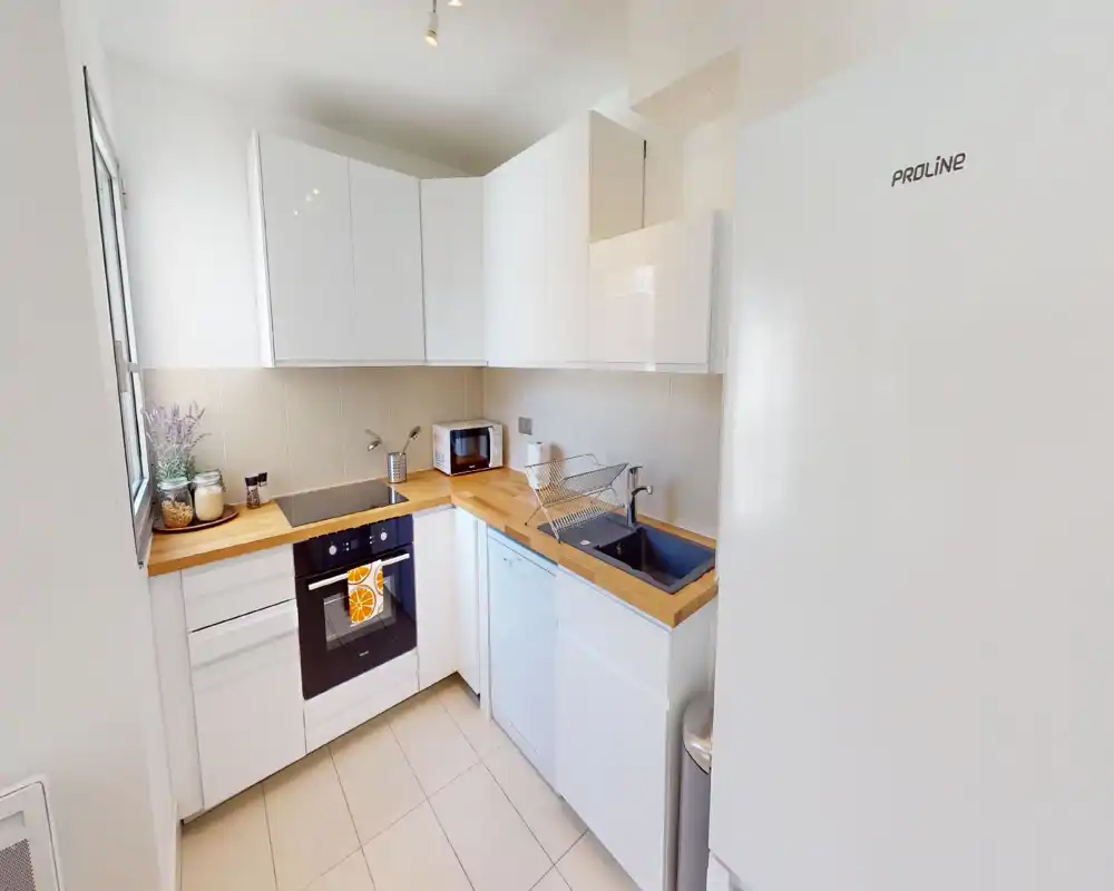 Compact modern kitchen with white cabinets, wooden countertops and integrated appliances. Bright, well organized space with a sink, oven, dishwasher and a small microwave; decorative jars and a plant add a cozy touch.