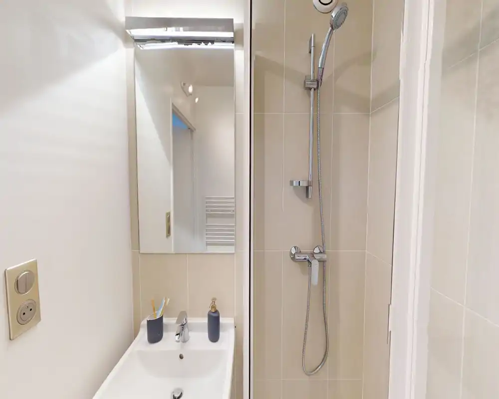 Compact modern bathroom with a wall-mounted shower and a small sink; light neutral tiles and chrome fittings create a clean, minimal look.