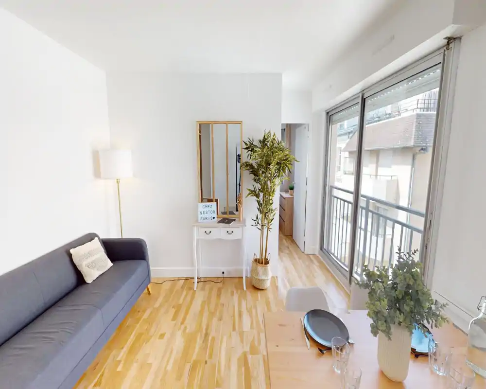 Bright, modern living room with a gray sofa, floor lamp and large sliding windows that open to a balcony. The space features warm wooden flooring, a small decorative console with mirror and plants, and a dining table visible in the foreground.