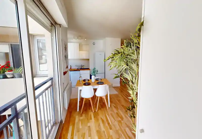 Bright, modern living area with a small dining table, two chairs and an open kitchenette; large sliding balcony door brings ample natural light and a pleasant wooden floor.