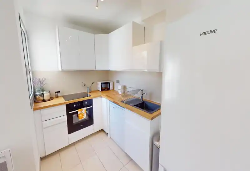 Compact modern kitchen with white cabinets, wooden countertops and integrated appliances. Bright, well organized space with a sink, oven, dishwasher and a small microwave; decorative jars and a plant add a cozy touch.