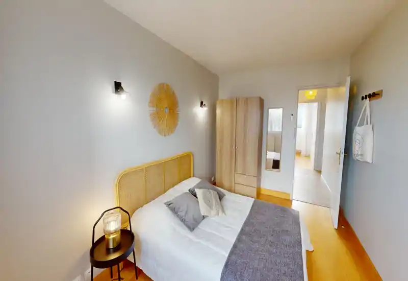 Camera da letto luminosa e moderna con testiera in rattan, letto matrimoniale rifatto, lampada da comodino e armadio. Pareti neutre e pavimento in legno caldo creano un'atmosfera accogliente, perfetta per annunci.