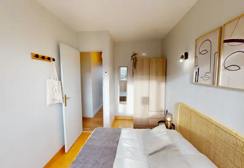 Chambre lumineuse et cosy avec tête de lit en rotin, linge de lit neutre et tableaux modernes épurés. La pièce dispose d'une armoire, d'une table de chevet avec lampe et d'un parquet chaleureux — parfaite pour une annonce locative.