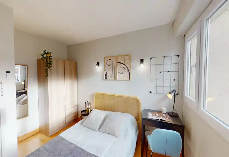 Chambre lumineuse et élégante avec tête de lit en rotin, table de chevet et armoire. Une grande fenêtre apporte la lumière naturelle et un petit bureau offre un espace de travail pratique.