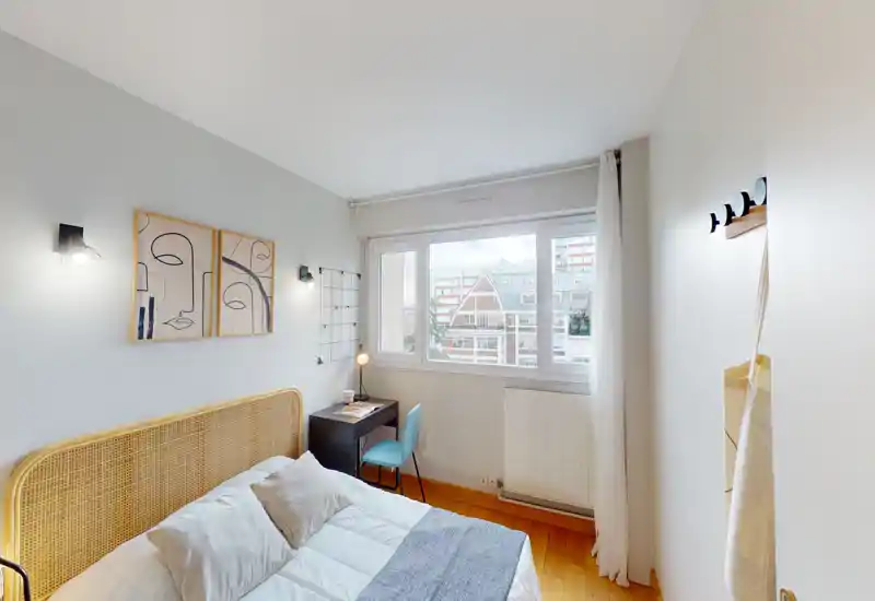 Chambre lumineuse et tendance avec tête de lit en rotin, lit bien fait et petit bureau près de la fenêtre offrant une vue sur la ville.