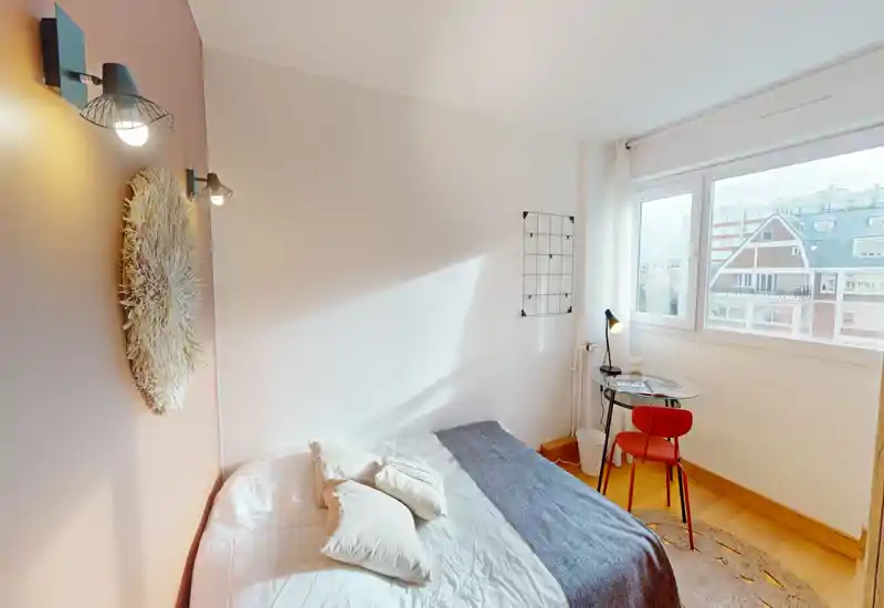 Chambre lumineuse et chaleureuse avec grande fenêtre, lit double habillé de linge de lit superposé et petit bureau près de la fenêtre — idéal pour travailler à distance.