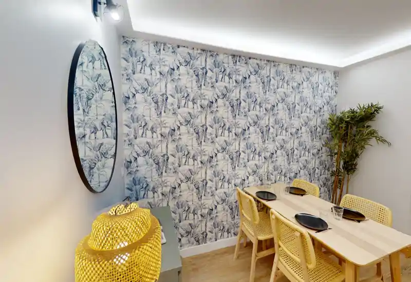 Zona pranzo elegante con tavolo in legno chiaro e sedie intrecciate, carta da parati decorativa e illuminazione calda—ideale per pranzi in piccolo gruppo.