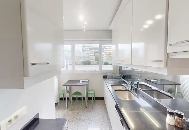 Cuisine moderne et lumineuse avec des placards blancs brillants, un long plan de travail noir avec double évier et une petite table avec tabourets verts près de la fenêtre.