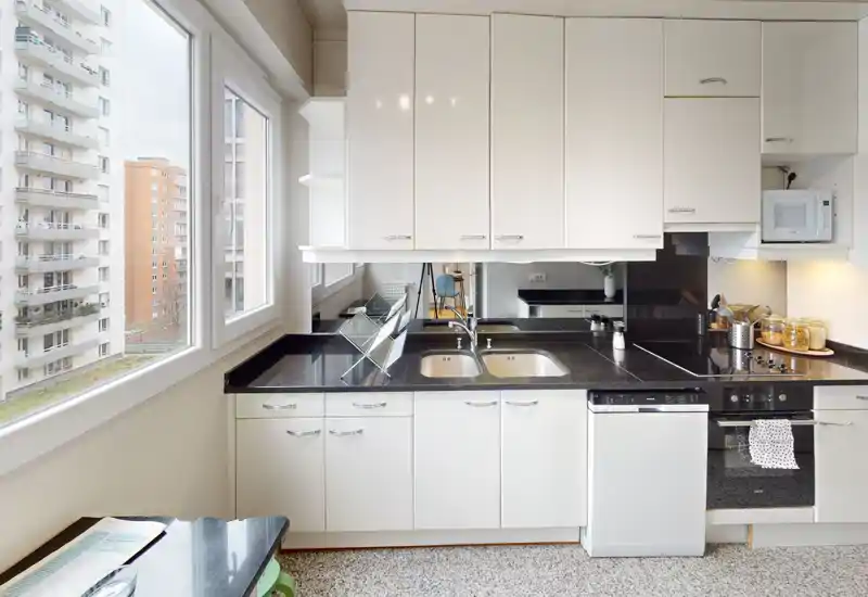 Cuisine moderne et lumineuse avec placards blancs, plan de travail noir, double évier et appareils intégrés ; grande fenêtre avec vue urbaine.