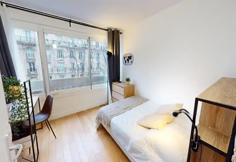 Bright, neatly staged bedroom with a comfortable single bed, bedside drawer, desk by a large window and warm lighting — ideal for a restful stay.