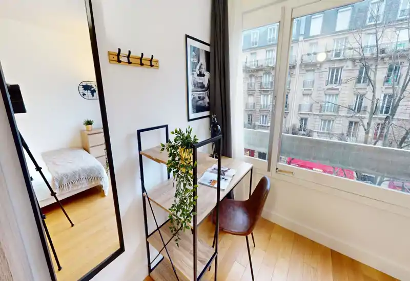 Sunny, compact bedroom with a visible single bed in an adjoining alcove, a stylish wooden desk by a large window and warm wood flooring — ideal for a student or single occupant.