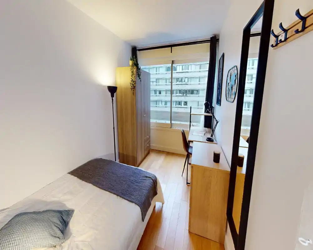 Dormitorio individual luminoso y bien decorado con cama cómoda, suelo de madera, armario y escritorio junto a una gran ventana que aporta mucha luz natural.
