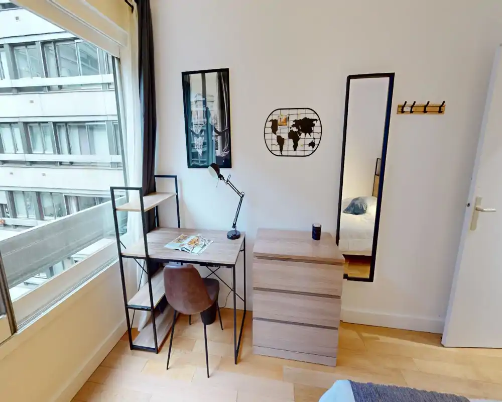 Rincón de dormitorio luminoso y moderno con escritorio y estantería junto a una gran ventana. Se aprecia una cómoda, una zona de trabajo con lámpara y silla y un atisbo de la cama reflejado en el espejo.