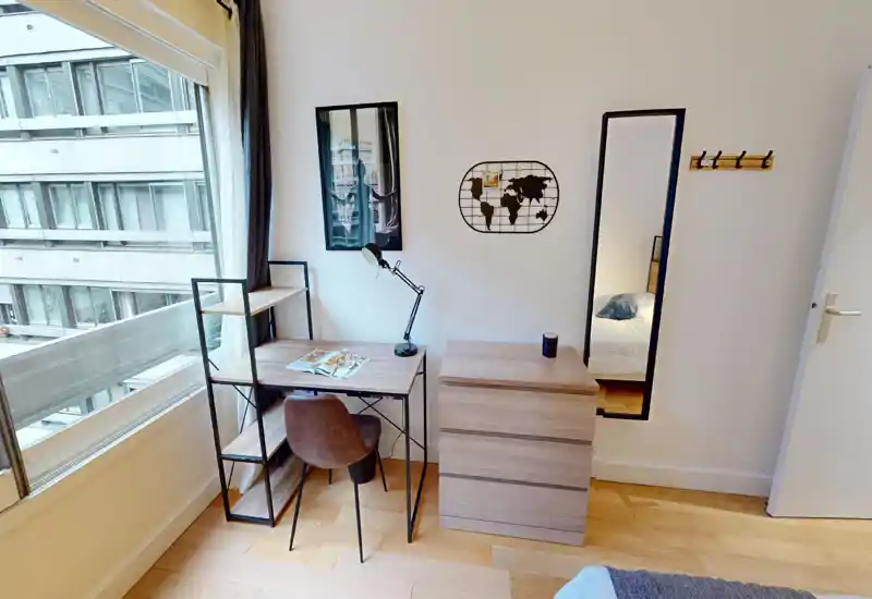 Rincón de dormitorio luminoso y moderno con escritorio y estantería junto a una gran ventana. Se aprecia una cómoda, una zona de trabajo con lámpara y silla y un atisbo de la cama reflejado en el espejo.