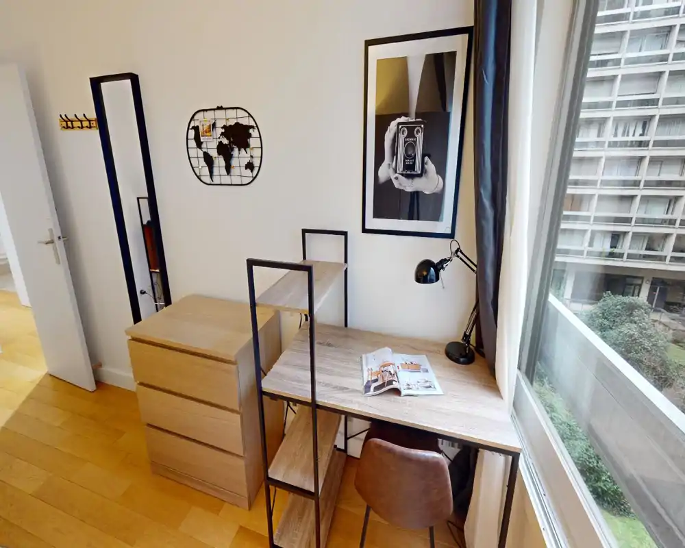 Bright, stylish living/working corner with a wooden desk, chair and chest of drawers by a large window. The space features framed art, a desk lamp and warm wood floors, ideal for remote work or study.