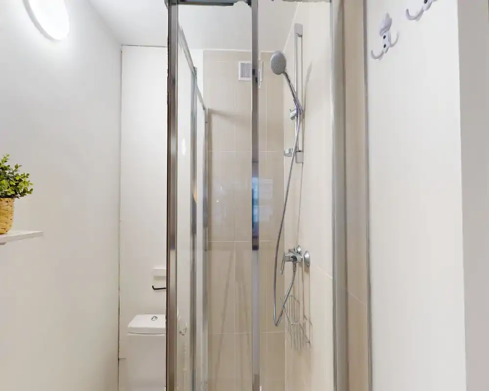 Bagno compatto e moderno con doccia piastrellata, soffione a mano e wc visibile; tonalità neutre e arredi puliti.
