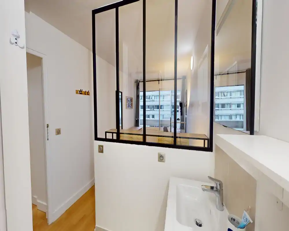 Angolo bagno compatto con lavabo bianco e miscelatore cromato moderno, separato dall'area giorno adiacente da una parete vetrata con cornice nera. Superfici pulite e luminose e alcuni prodotti da toiletta visibili suggeriscono una soluzione en-suite pratica collegata alla stanza principale.