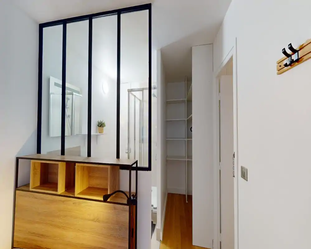 Bagno moderno e luminoso con box doccia in vetro, mobile specchiato e pareti bianche pulite. Pavimento in legno visibile all’esterno e scaffalature aperte nell’ingresso.