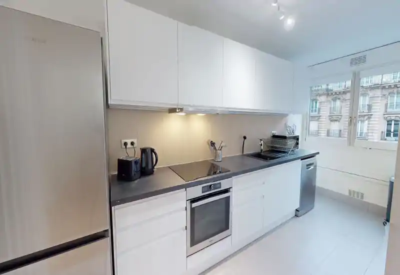 Bright modern kitchen with white cabinetry, integrated oven and induction hob, stainless fridge and dishwasher, and a large window providing natural light.