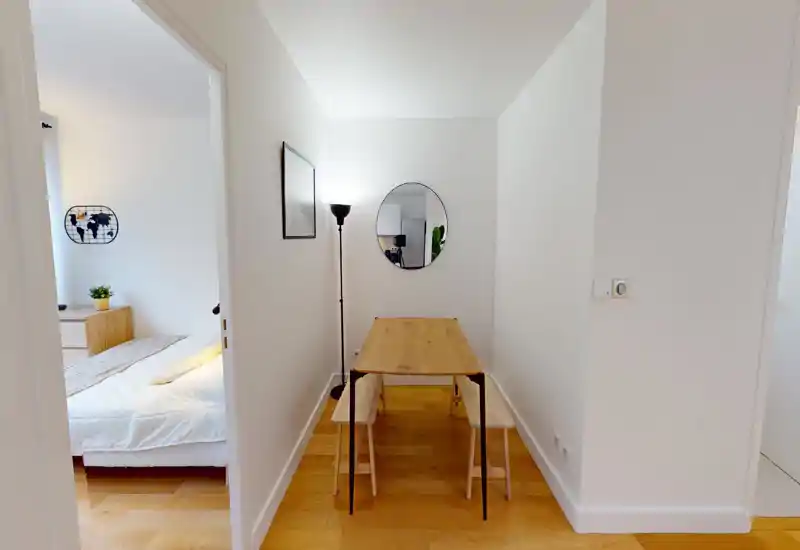 Compact dining nook with a minimalist wooden table and benches, a standing lamp and a round wall mirror that visually expands the space. Bright, clean styling with warm wooden floors.