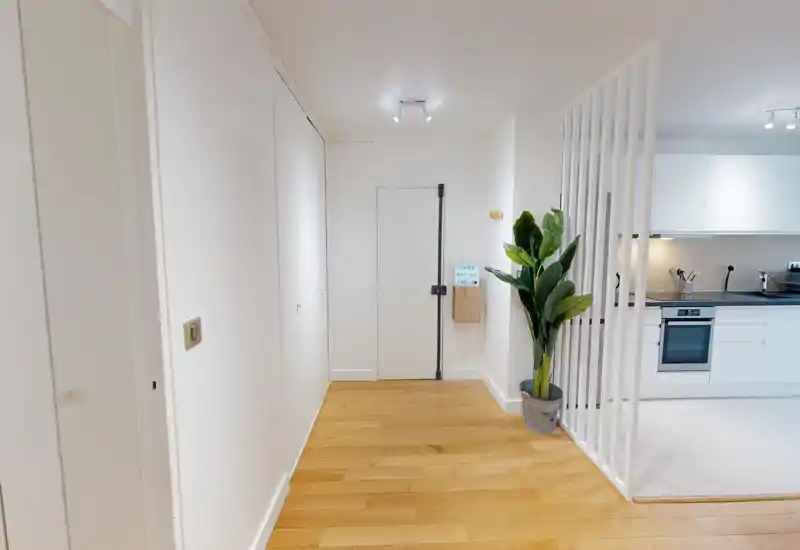 Bright, minimal entrance hallway with light wood flooring, a white front door and a decorative potted plant; the kitchen is visible through a slatted divider.