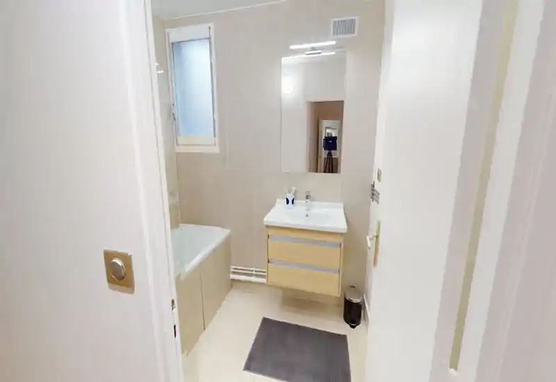 Clean, modern bathroom with a built-in bathtub, wall-mounted sink cabinet and a large mirror. Neutral tiles and good lighting create a bright, tidy space ideal for rental listings.