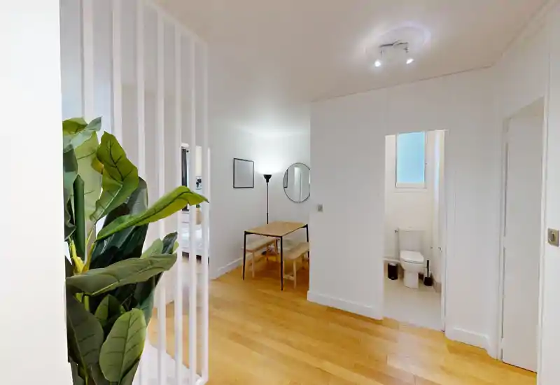 Zona de estar luminosa y moderna con suelo de madera, mesa pequeña con bancos y plantas decorativas; diseño abierto que conecta con dormitorio y baño compacto.