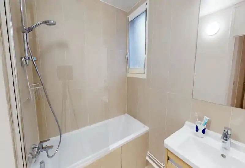 Clean, modern bathroom featuring a bathtub with wall-mounted shower, tiled walls, a frosted window and a sink with mirror — bright and functional.