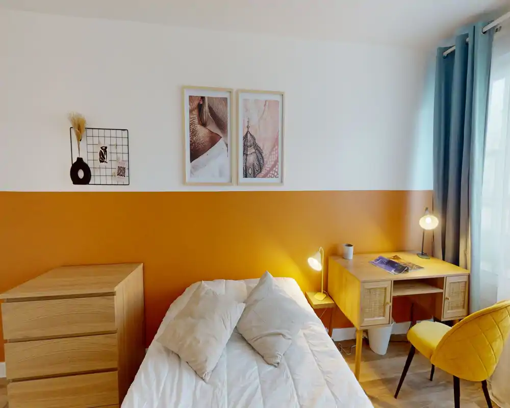 Chambre lumineuse et stylée avec lit bien fait, mur d'accent ocre, commode en bois et petit bureau près de la fenêtre — idéale pour se reposer et travailler légèrement.