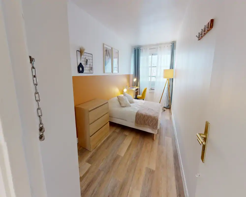 Chambre lumineuse et décorée avec lit double, commode et bureau près de la fenêtre; mur accent ocre et sol imitation bois pour une ambiance chaleureuse et moderne.