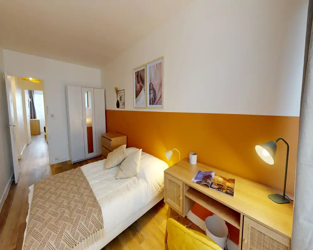Chambre lumineuse et soignée avec lit double, lampes de chevet, bureau et armoire; mur accent ocre et sol en bois pour une ambiance chaleureuse.