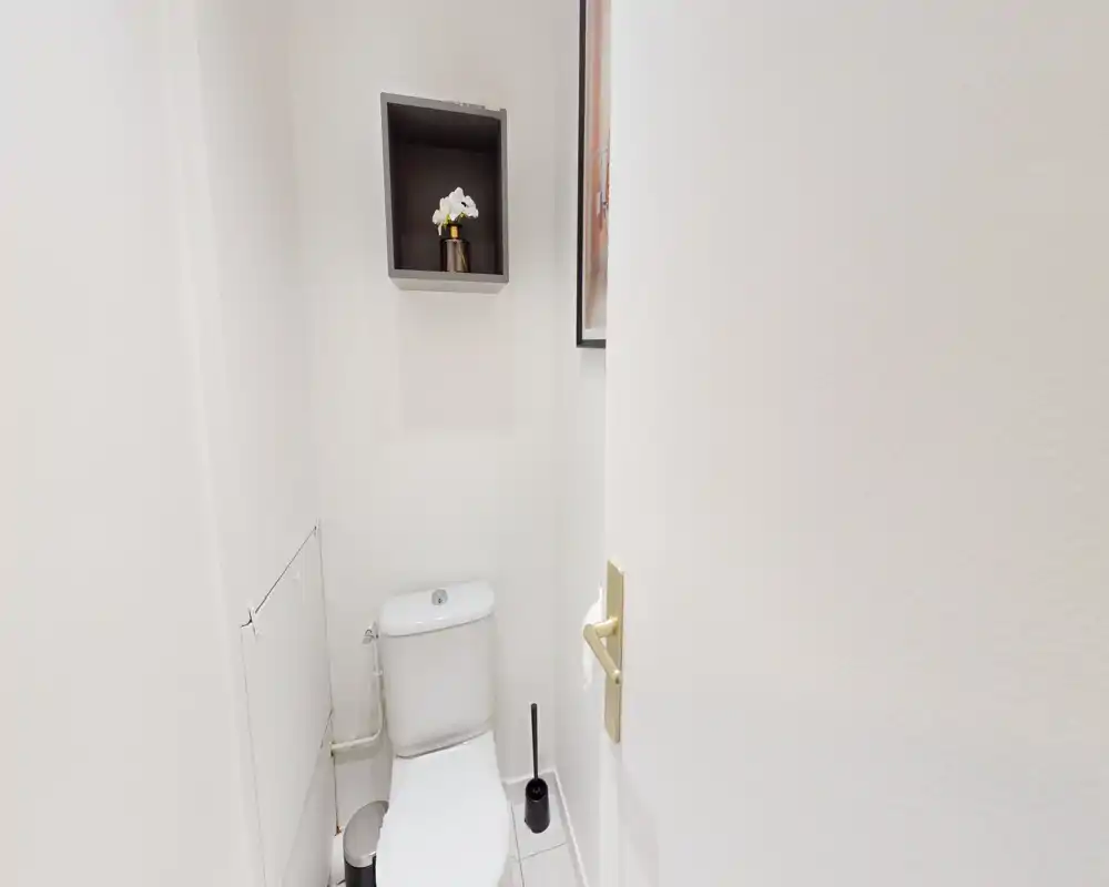 Compact private toilet with a modern white cistern, tiled floor and simple decorative shelf above; clean and well-lit.