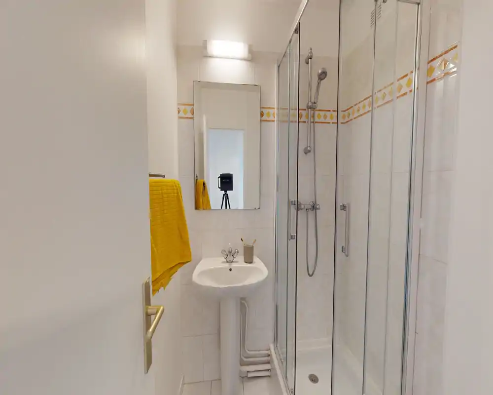 Compact, clean bathroom with corner glass shower, pedestal sink and a wall mirror. White tiles and a yellow towel add a fresh touch.