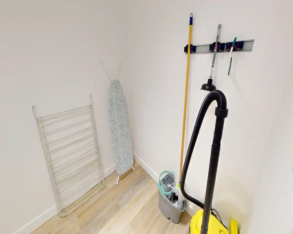 Small utility/storage room with cleaning tools—vacuum cleaner, mop bucket, ironing board and drying rack on a light wood floor; bright, functional space suitable for laundry or storage.
