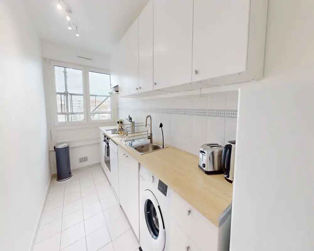 Bright, narrow galley kitchen with white cabinets, wooden countertop, sink, washing machine and small appliances; clean, modern and efficiently arranged for compact urban living.