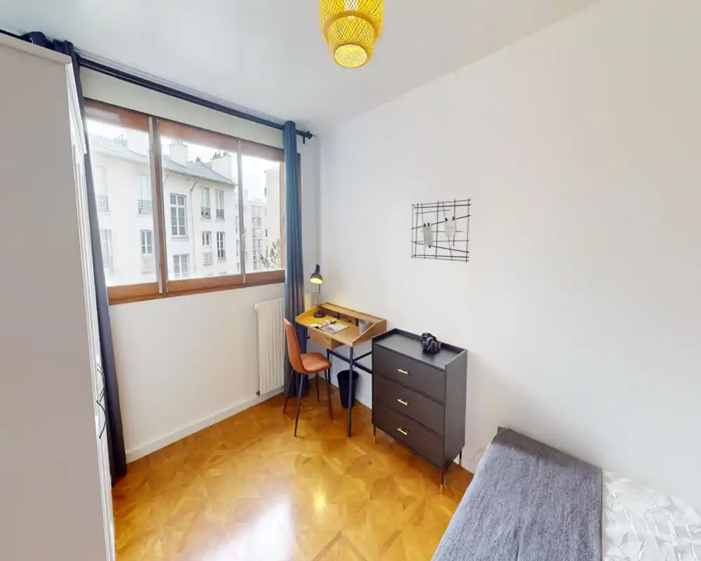 Chambre lumineuse et soignée avec un lit simple visible à droite, un petit bureau en bois avec lampe près de la fenêtre et une commode à trois tiroirs. Grande fenêtre apportant de la lumière naturelle et parquet chaleureux.
