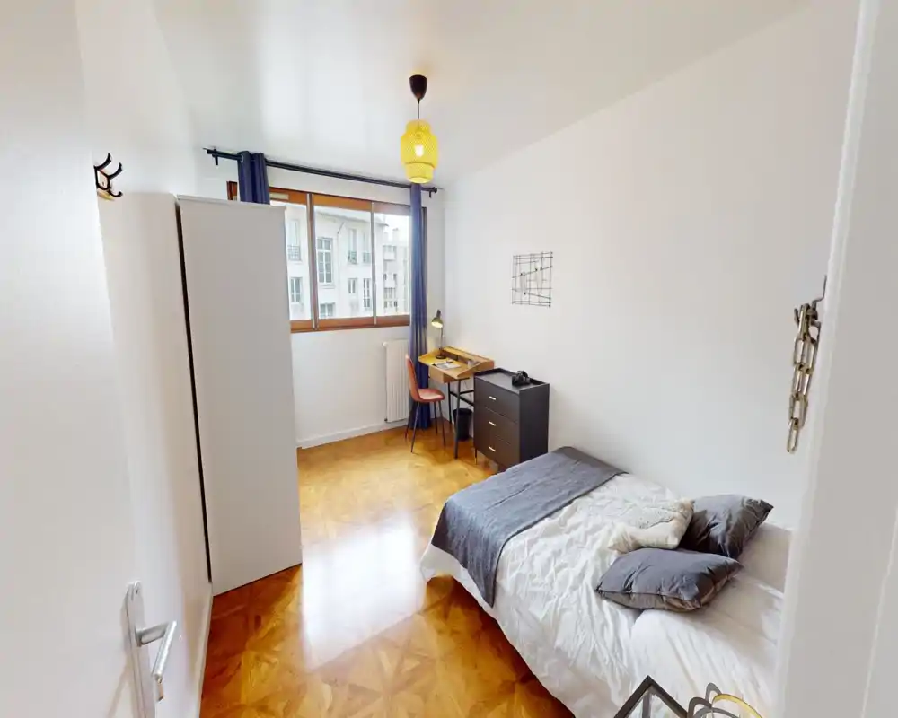 Chambre lumineuse et soignée avec un lit simple, parquet en bois et un petit bureau près de la fenêtre — idéale pour un étudiant ou une personne seule.