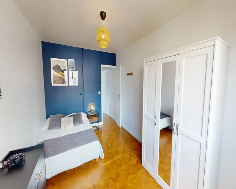 Chambre lumineuse et soignée avec un lit double contre un mur d'accent bleu, une table de chevet avec lampe et une grande armoire blanche avec miroir. Parquets cirés et décor minimaliste pour une atmosphère propre et accueillante.