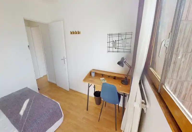 Dormitorio luminoso y ordenado con cama individual y escritorio de madera junto a la ventana — ideal para estudiantes o profesionales. La luz natural llena la habitación y el escritorio ofrece un espacio de trabajo funcional.