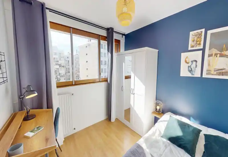 Dormitorio luminoso y decorado con cama visible, pared de acento azul verdoso, armario con espejo y escritorio de madera junto a la ventana. Ventana amplia que aporta mucha luz natural, cuadros y textiles que crean un ambiente cuidado.