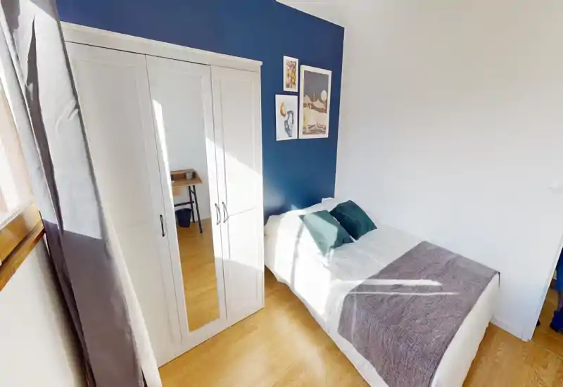 Dormitorio moderno y luminoso con cama doble junto a una pared azul oscuro, cojines decorativos y una manta. Armario blanco con puerta espejo y suelo de madera completan el espacio minimalista y soleado.