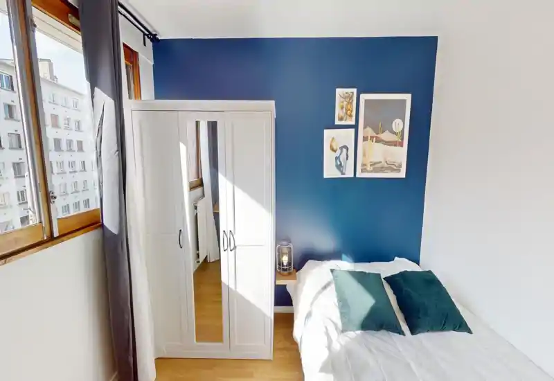 Dormitorio luminoso con pared azul, armario blanco con puerta espejo y cama bien hecha con cojines verdes; ideal para anuncios que destaquen la luz natural y la decoración moderna.